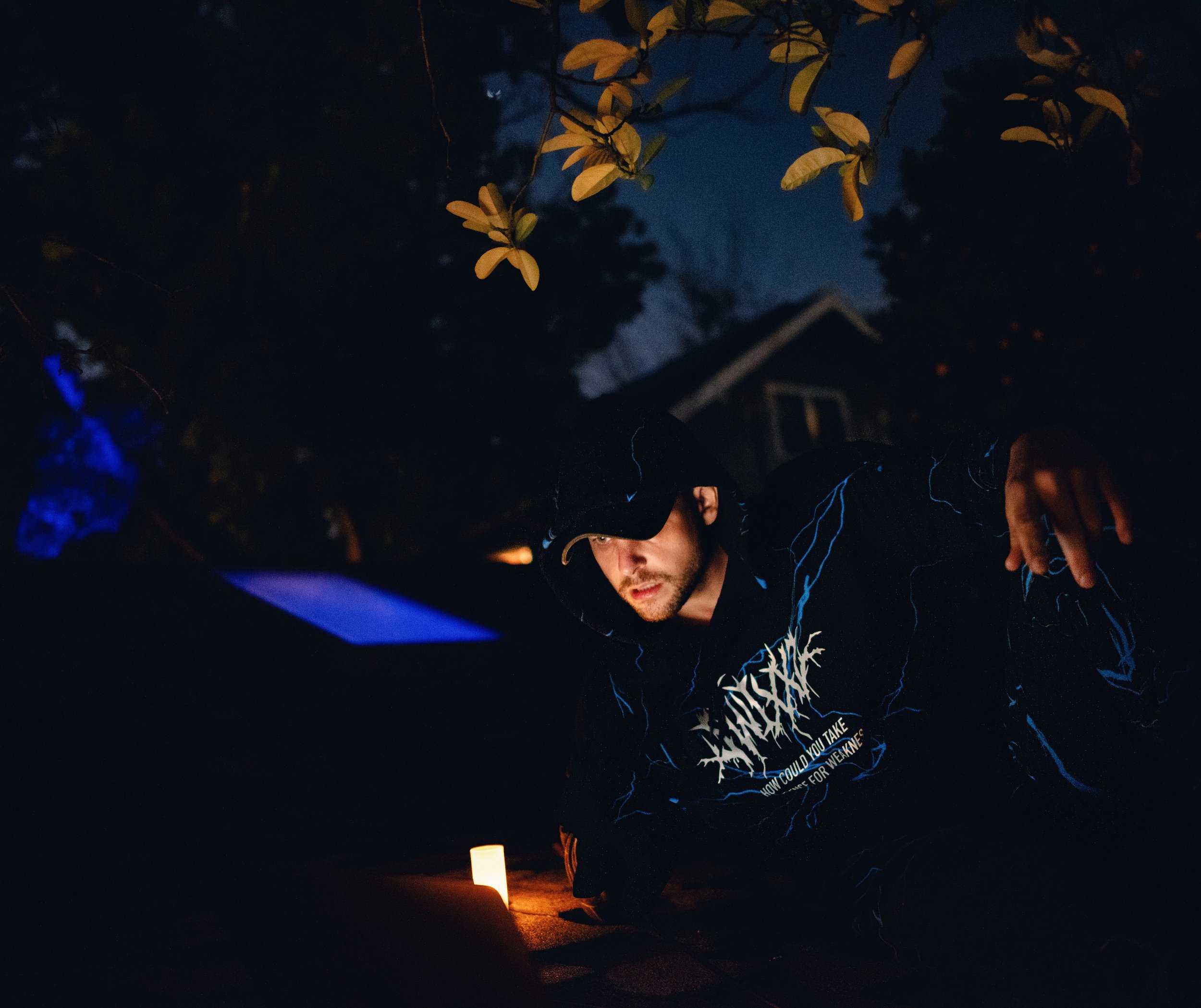 Cody Tarpley photographed at night in a hooded jacket, lit by a single candle on the pavement.